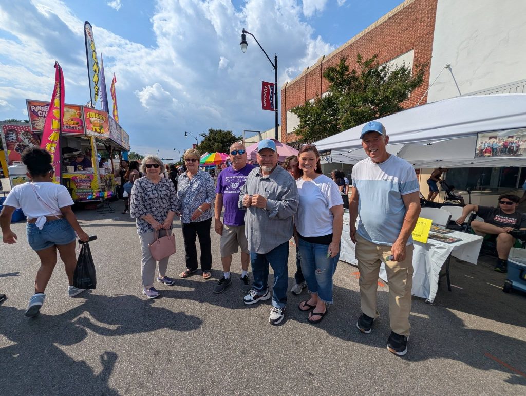 A group of people poses together at an outdoor street festival, with food trucks, vendor booths, and other festival-goers in the background under a partly cloudy sky.
