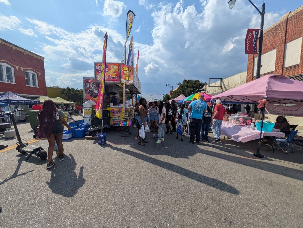 People gather at a street fair with food trucks, vendor tents, colorful umbrellas, and a bright sky filled with clouds overhead.