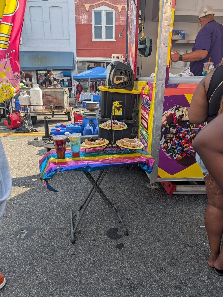 Colorful table with waffles topped with fruit and powdered sugar, three drinks, in front of a food truck at a lively outdoor street market.