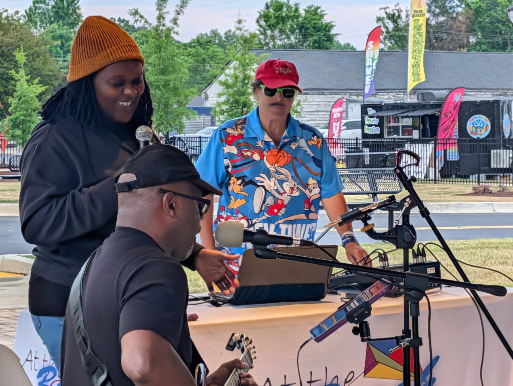 Three people performing music outdoors, one singing, one playing guitar, and one at a table with equipment. Colorful banners and food trucks are visible in the background.
