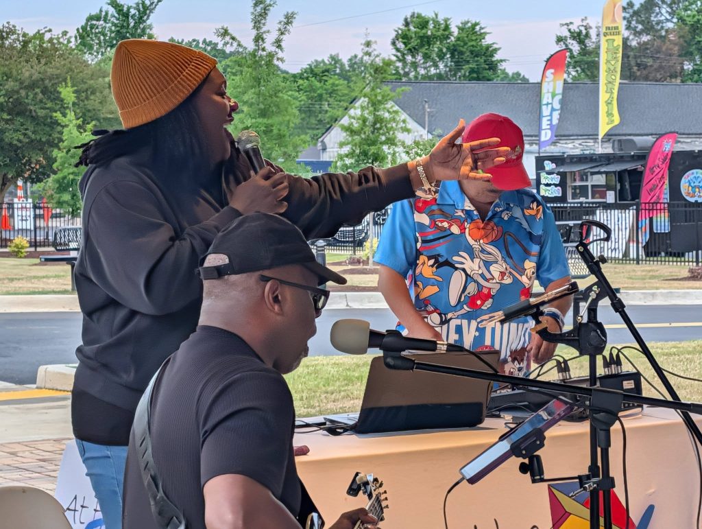 Three musicians performing outdoors, one singing with a microphone, one playing guitar, and one using DJ equipment. Bright, colorful banners and trees visible in the background.