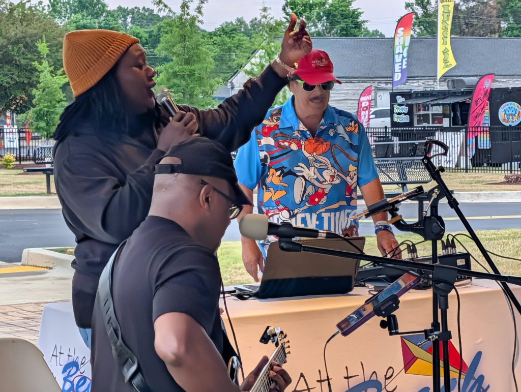 Three musicians perform outdoors; one sings, another plays guitar, and a third operates a laptop. Banners and a food truck are visible in the background.