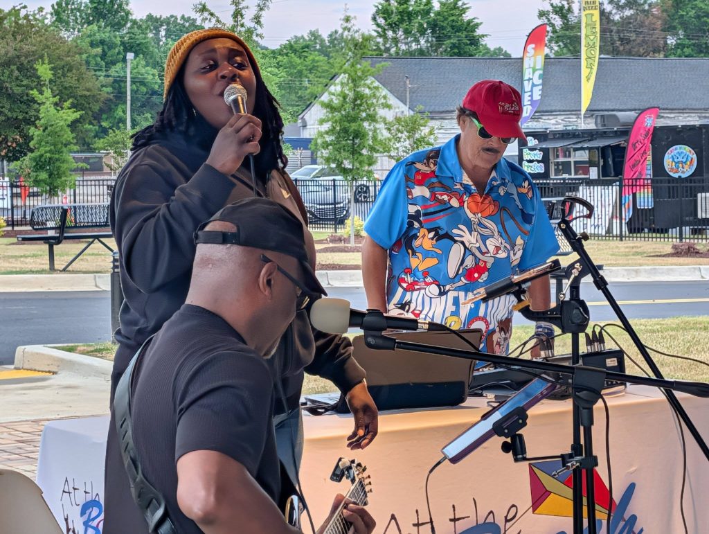 Three musicians performing outdoors: a woman singing, a man playing guitar, and another man at a DJ setup wearing a cartoon shirt and red cap.