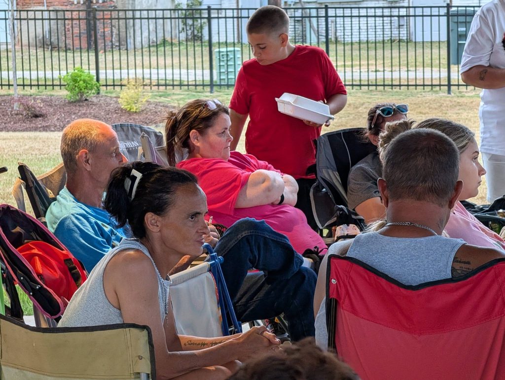 Several adults and a child gather outdoors, sitting in camping chairs. The child holds a food container. The setting appears relaxed and social, with greenery and a fence in the background.