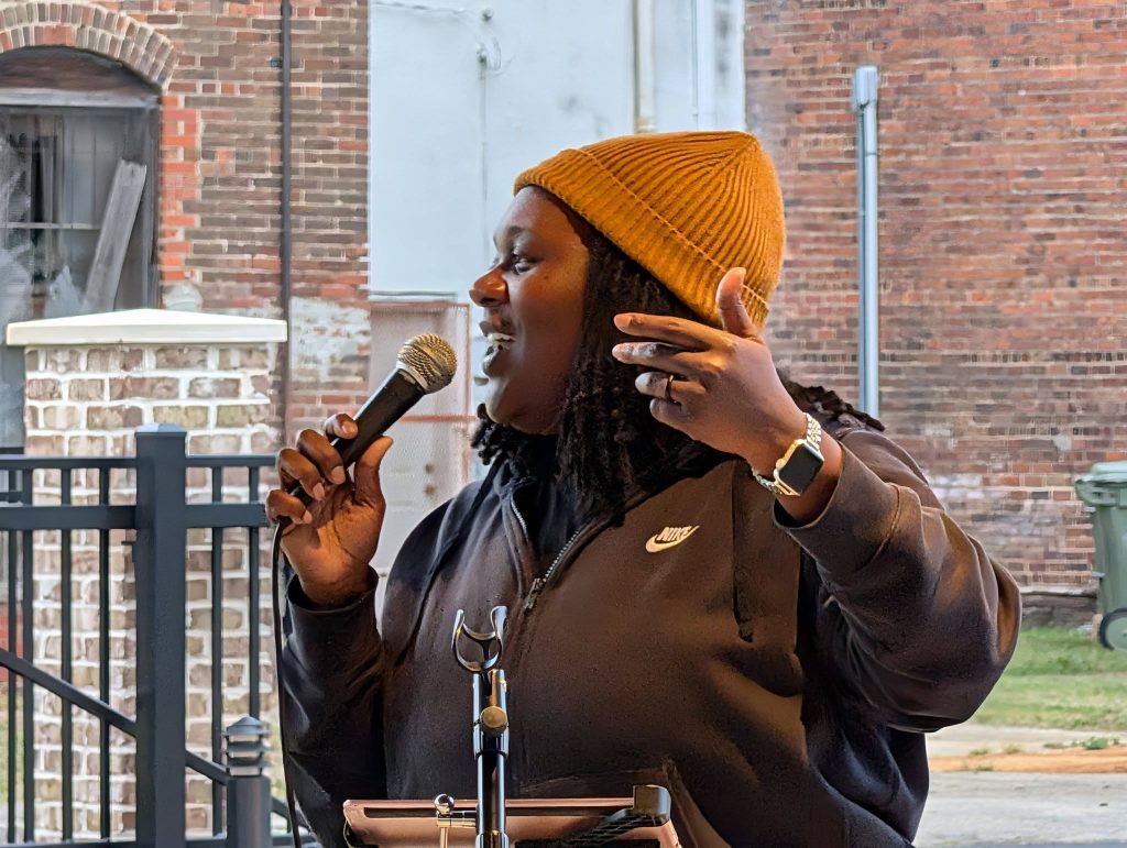 Person wearing a mustard beanie and black jacket speaks into a microphone outside, with a brick wall and iron fence in the background.