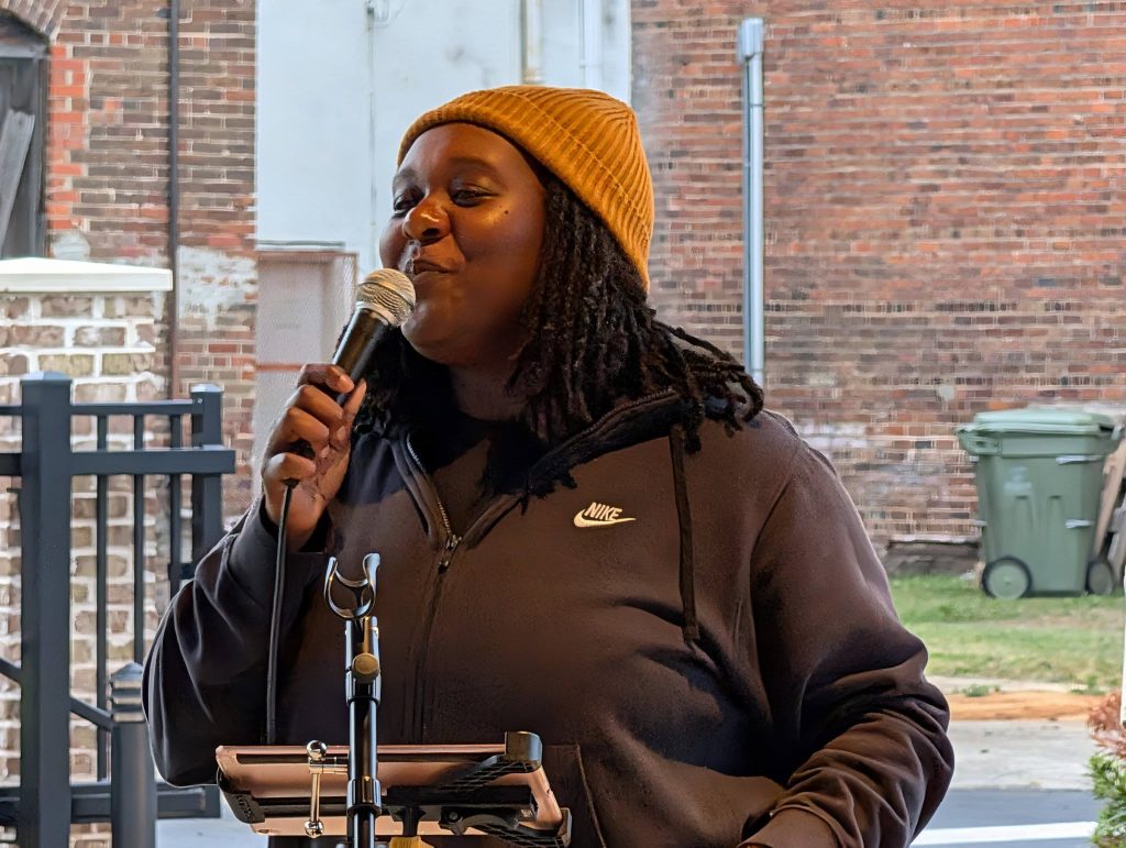 Person wearing a brown beanie and black Nike hoodie speaks into a microphone outdoors, standing in front of a brick wall and some fencing.