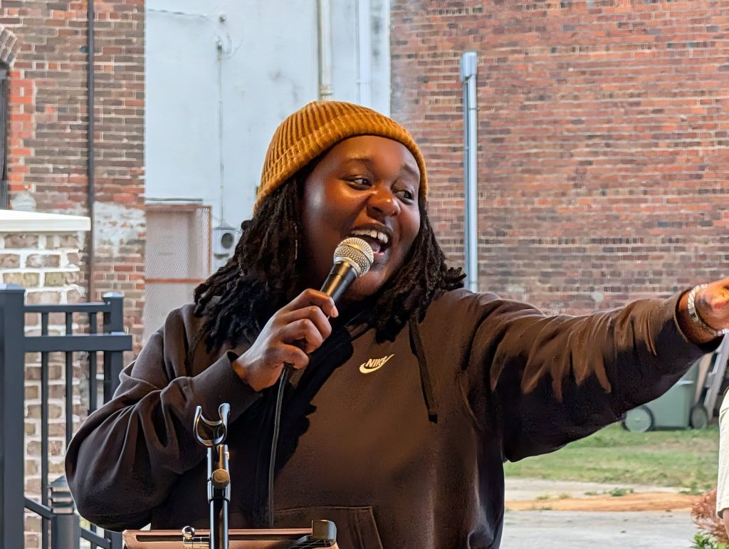 Person wearing a brown beanie and black hoodie speaks into a microphone and points, standing outdoors in front of a brick wall.