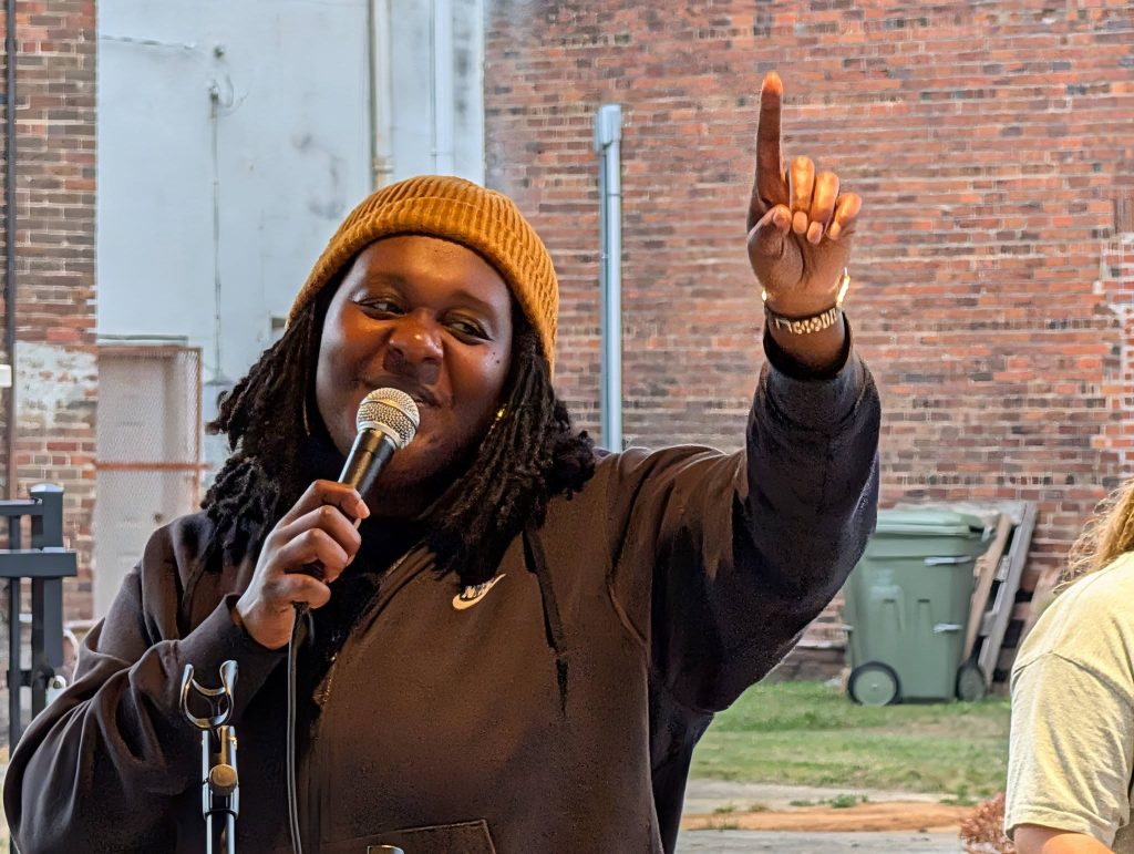 Person in brown beanie and black jacket speaking into a microphone, smiling and pointing upward, standing outdoors in front of a brick wall.