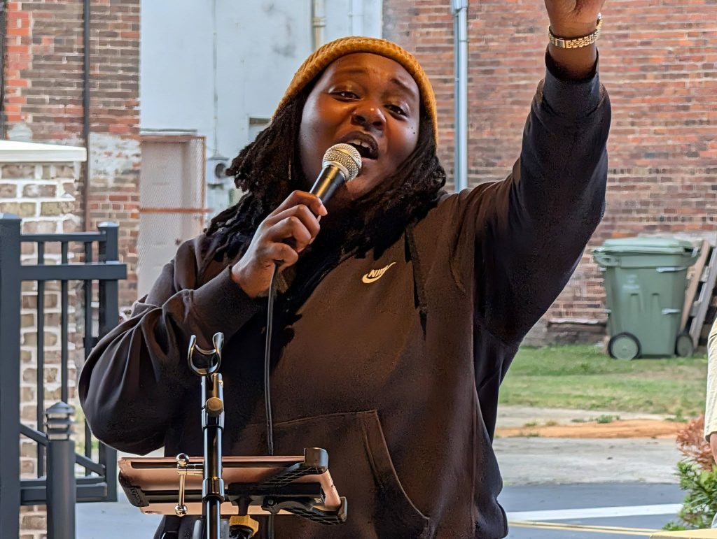 Person wearing a brown hoodie and yellow beanie sings passionately into a microphone outdoors, raising one arm. Brick wall and trash bins visible in the background.