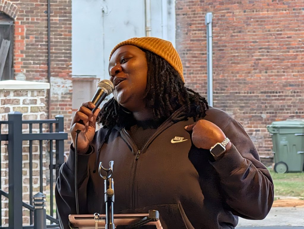 Person wearing a brown beanie and black Nike hoodie speaks into a microphone outdoors, standing in front of a brick wall and metal gate.