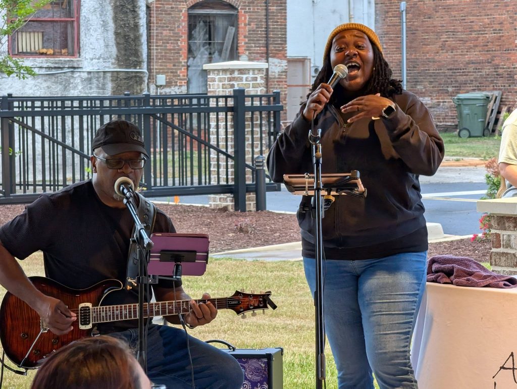 A man playing electric guitar and a woman singing into a microphone perform outdoors, with a gate and brick buildings in the background.
