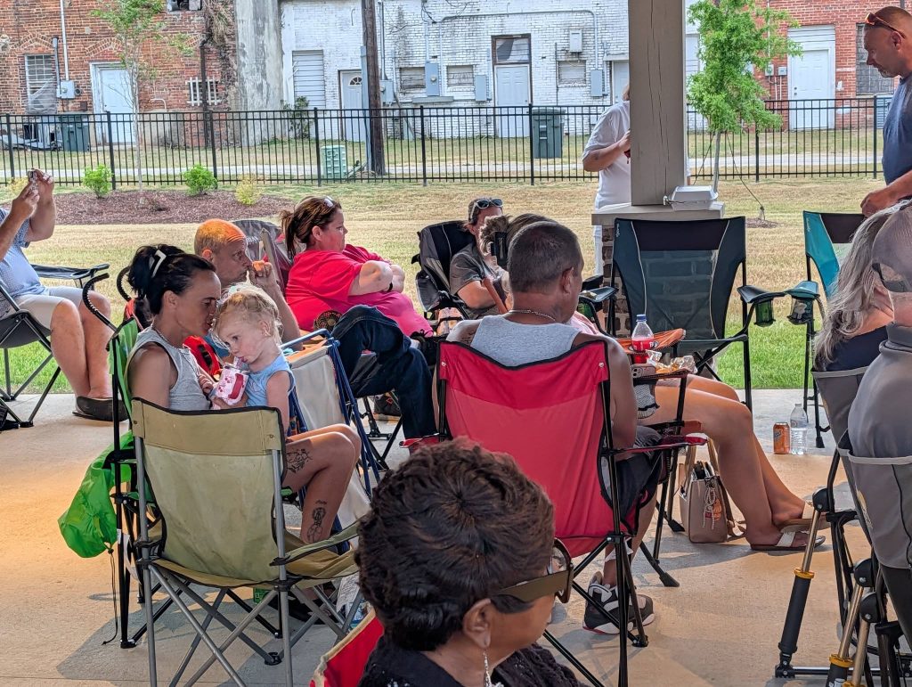 People sitting in folding chairs under a pavilion, some eating and talking, with a child drinking from a pouch. Urban background with buildings and grass outside.