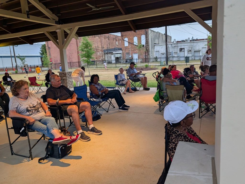 People seated in lawn chairs under a pavilion, some watching an event, with buildings and people visible outside in the background.