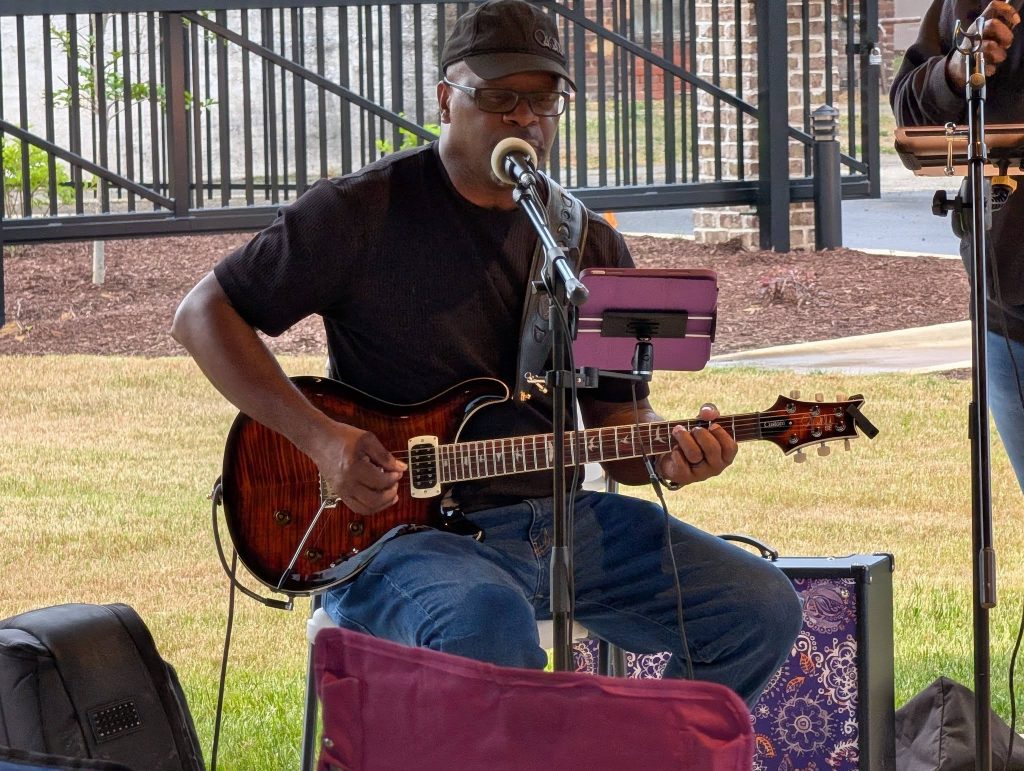 Man wearing a cap and glasses plays an electric guitar and sings into a microphone outdoors, seated under a shelter with music gear nearby.