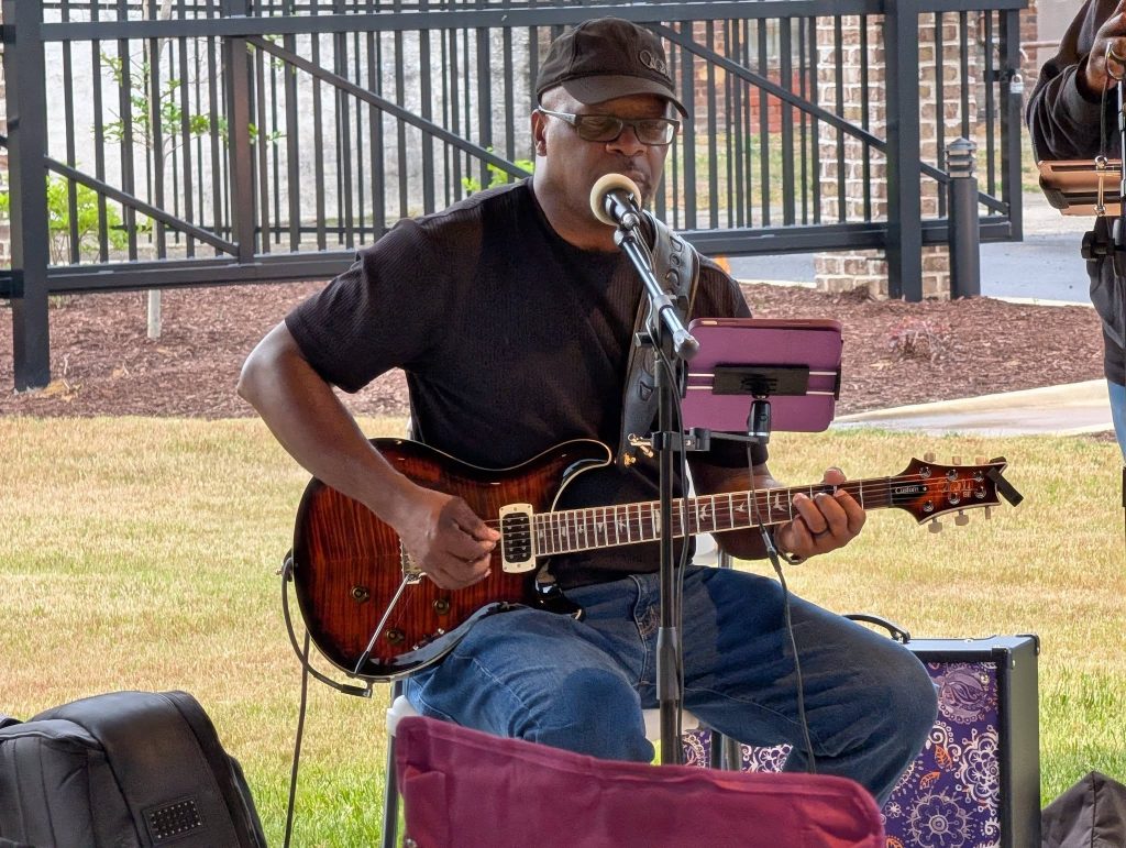 Man wearing a hat and glasses, playing electric guitar and singing into a microphone outside on a grassy area, with musical equipment nearby.