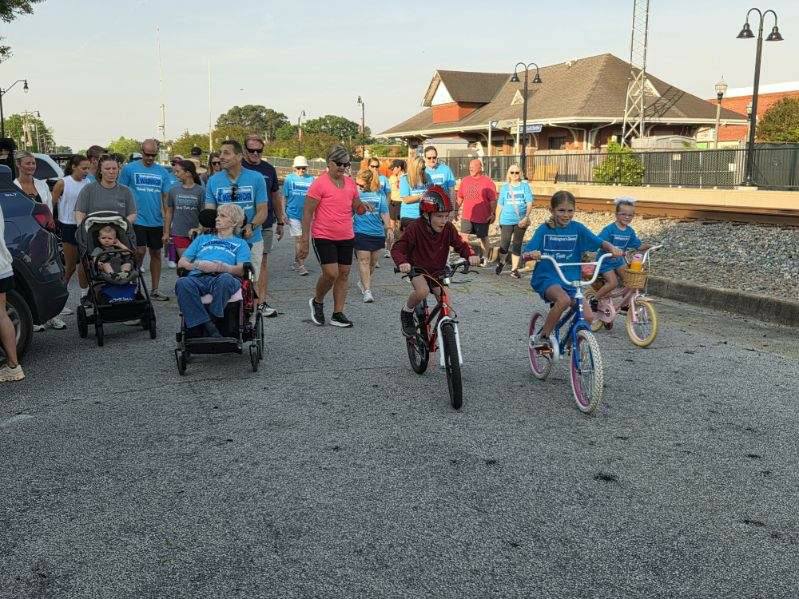 A group of people, including kids on bikes and individuals in wheelchairs, participate in a community walk near a railway station on a sunny day.