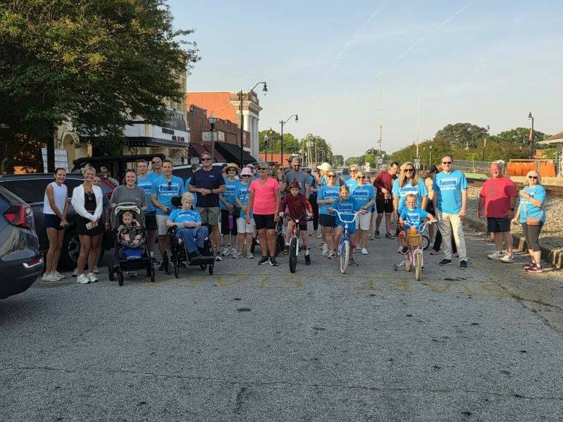 A group of people, including children on bikes and individuals in wheelchairs, gathered outdoors on a street for a community event on a sunny day.