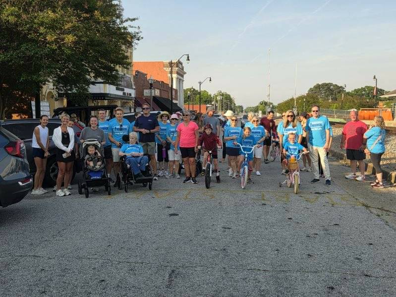 A group of people, including children on bikes and adults, gather on a street for a community event, many wearing matching blue T-shirts.