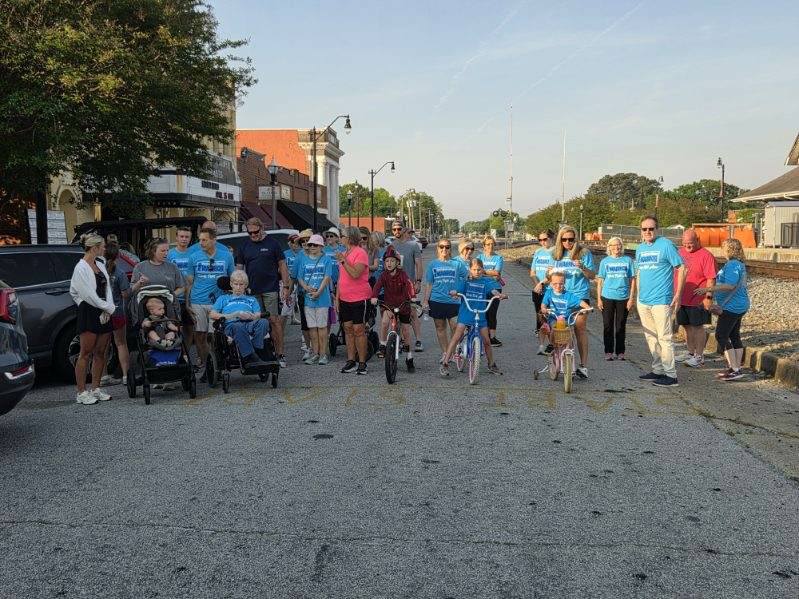 A group of people, including children on bikes and some in wheelchairs, gather on a city street wearing matching blue shirts for an event.