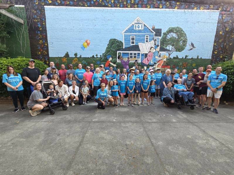 A large group of people pose together in front of a colorful mural featuring a house, children, and balloons painted on a brick wall.