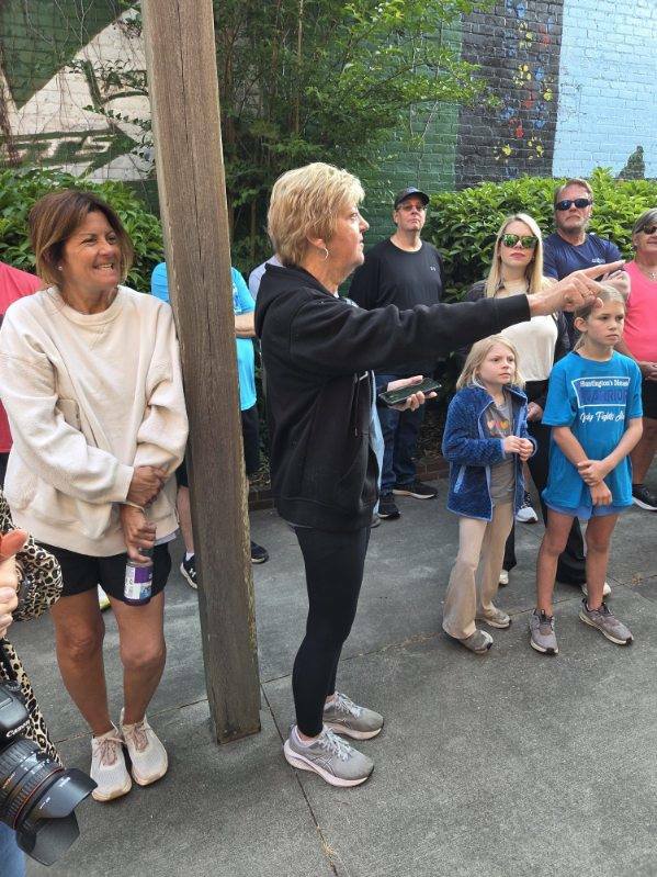 A group of people, including adults and children, stand outdoors. One woman in black points while others listen and observe, some dressed casually in shorts or athletic wear.