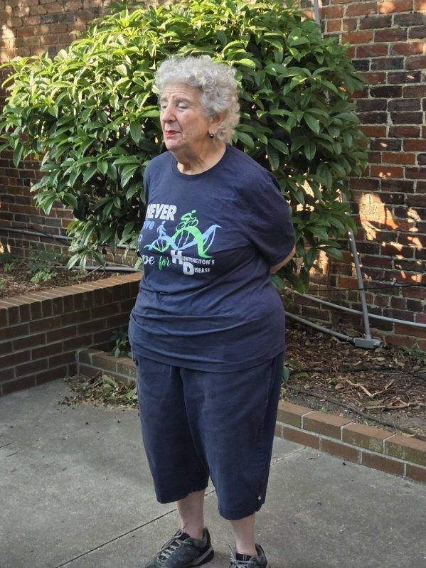 Elderly woman with curly hair stands outdoors in casual clothing, in front of a leafy shrub and brick wall, hands behind her back.