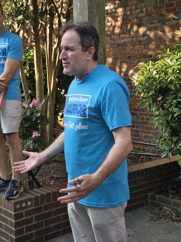Man in blue "Warrior" T-shirt speaking outdoors near brick wall and plants, gesturing with hands.