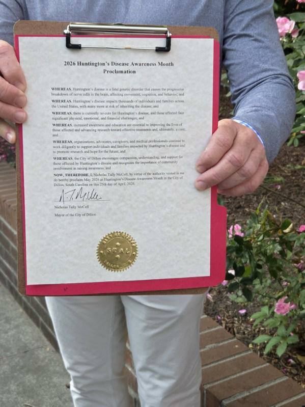 A person holds a clipboard displaying a 2026 Huntington’s Disease Awareness Month proclamation, featuring a gold seal, signature, and printed text, with flowers and a brick wall in the background.
