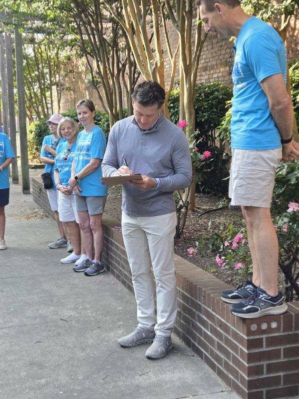 A man writes on a clipboard while people in blue shirts stand and watch near a garden area with brick walls and flowering shrubs.