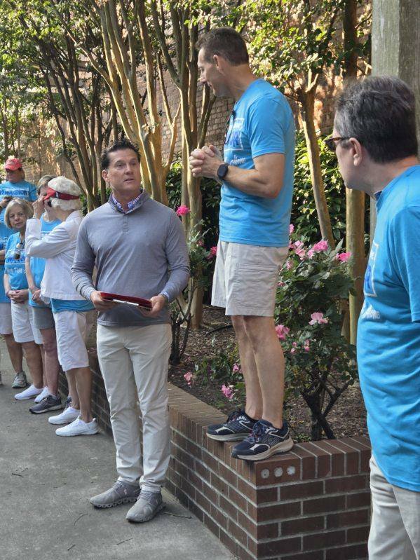 A group of people standing outdoors near trees and flowers, with one man standing on a low brick wall, engaging in conversation.