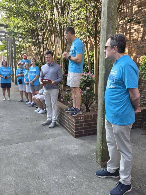 A group of people, some in matching blue shirts, stand outdoors near a brick wall while one man speaks and another holds a tablet.