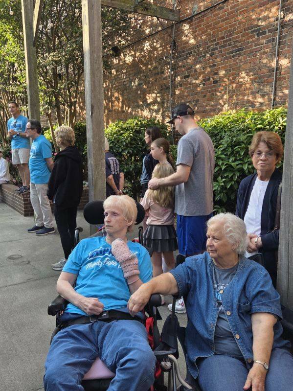 A group of people, including seniors and children, gather outdoors near a brick wall and greenery on a sunny day.