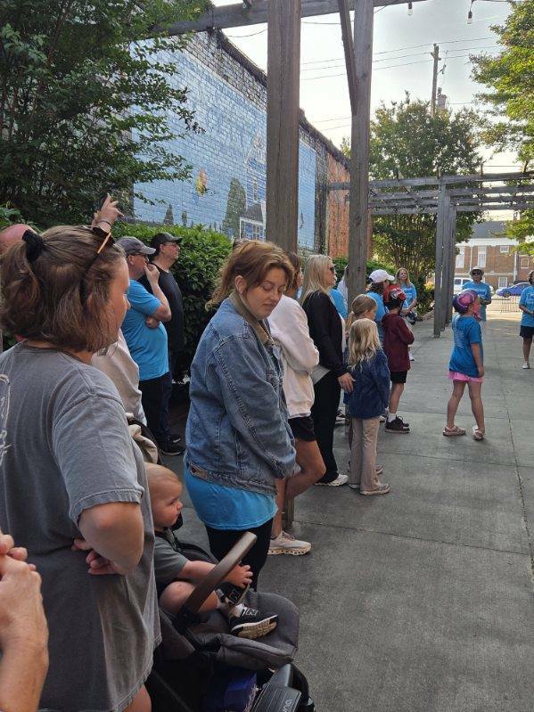 A group of people, including adults and children, gather outside near a mural and pergola, some wearing blue shirts, on a sunny day.