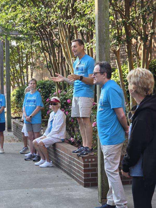 A group of people wearing blue shirts gather outdoors. One man stands on a brick ledge speaking to the group. Trees and flowers surround them.