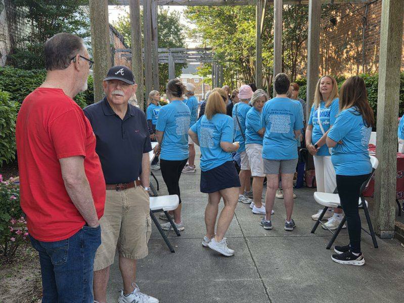 People gather outdoors, many wearing matching blue shirts, some standing and some sitting on white chairs, engaging in conversation under a wooden pergola structure.