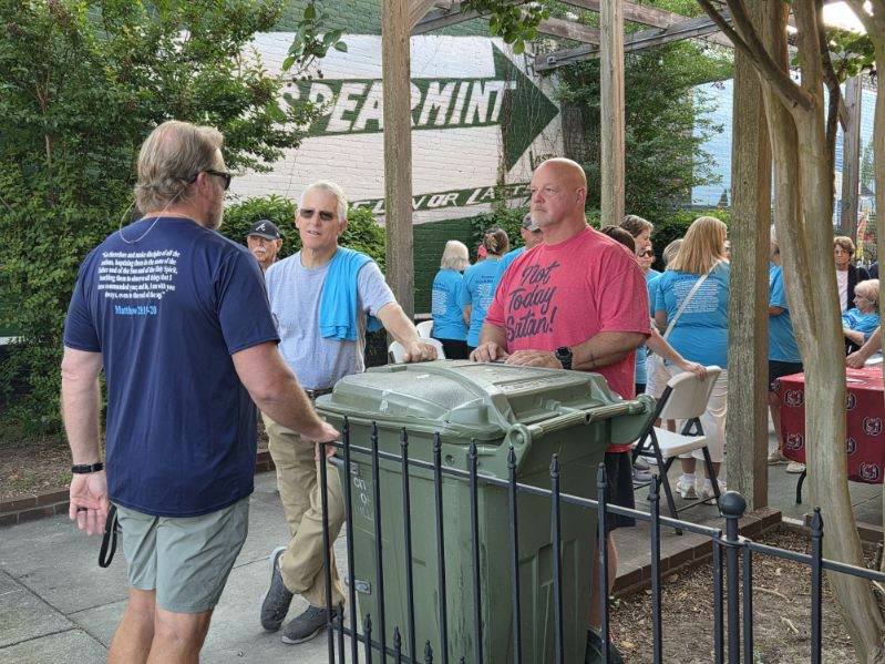 Three men talking near a trash bin outdoors; one wears a shirt reading "Not Today Satan!" while people in blue shirts gather in the background.