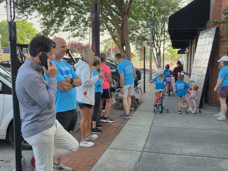 Adults and children in casual clothes gather on a sidewalk, some standing, some on bicycles, many wearing matching blue shirts, with trees and parked cars nearby.