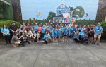 A large group of people pose together in front of a colorful mural depicting a blue house, trees, and children reading books.