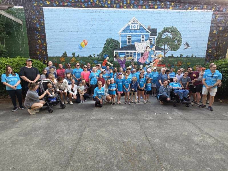 A large group of people pose together in front of a colorful mural depicting a blue house, trees, and children reading books.