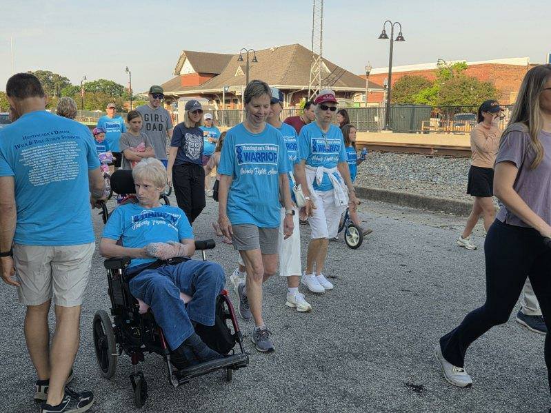 A group of people walking outdoors, many wearing blue "Warrior" shirts, with one person in a wheelchair, participating in what appears to be a community event.