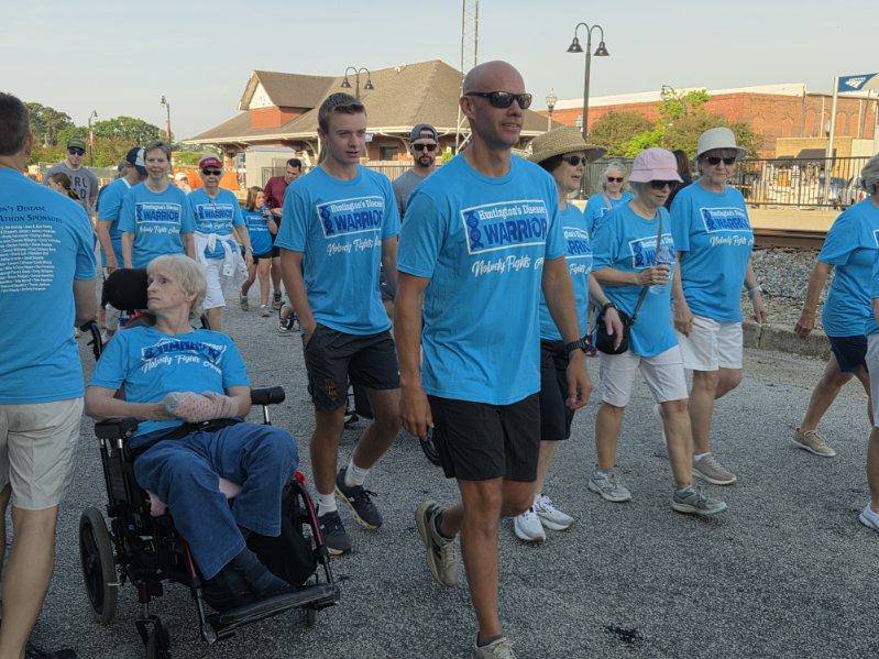 A group of people, some wearing blue "Warrior" T-shirts, walk outdoors together; one person is in a wheelchair, participating in the event.