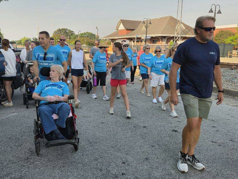 A group of people participate in an outdoor walk event, some wearing matching blue shirts, including one person in a wheelchair being pushed by another.