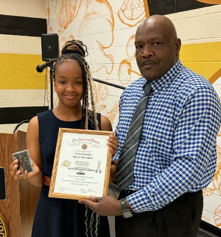 A young woman and a man pose together, with the woman holding a certificate and a small box, standing indoors against a decorated wall.