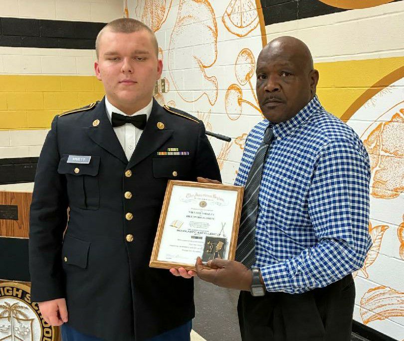 A young man in a formal military uniform stands next to an older man holding a framed certificate, both posing indoors against a decorated wall.