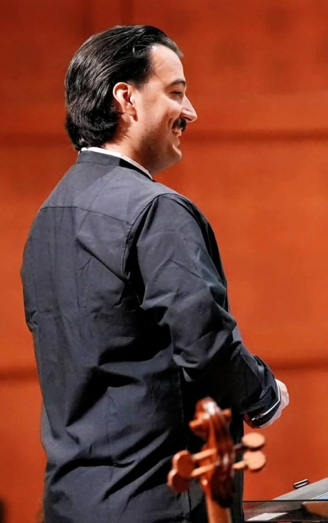 A man in formal attire stands on stage, smiling, with an orange-brown background and a musical instrument partially visible in the foreground.