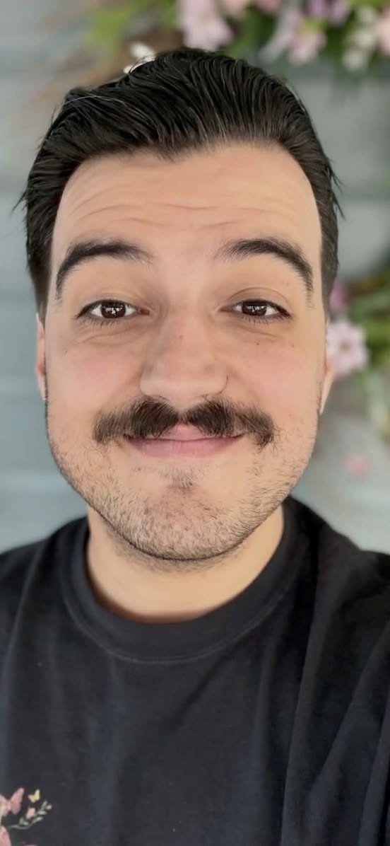 A man with dark hair, mustache, and stubble smiles at the camera while wearing a black shirt. Flowers and greenery are blurred in the background.