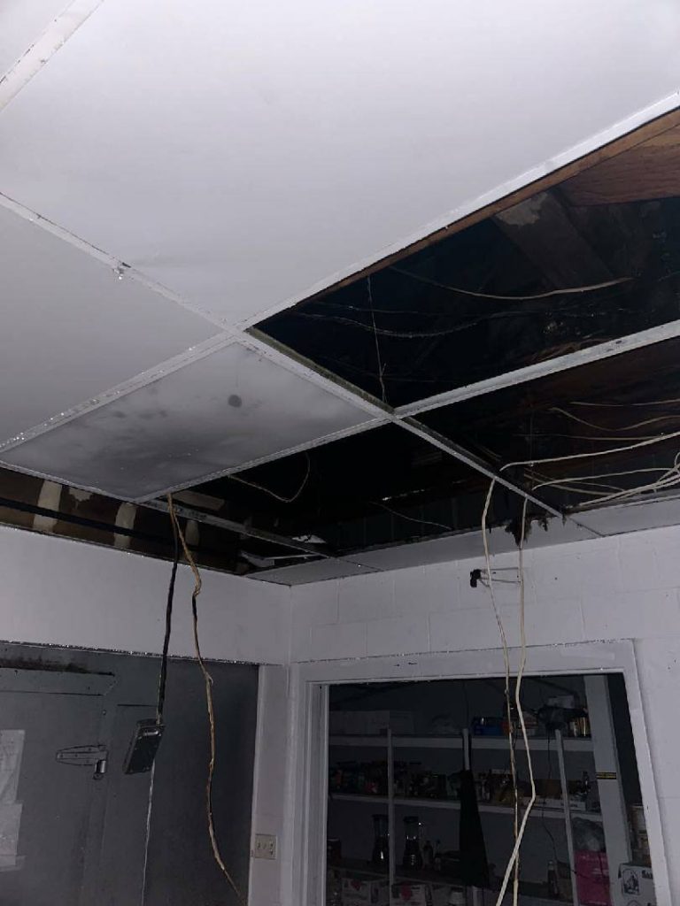 Ceiling with missing panels revealing exposed wires and beams above a room with shelves containing various household items.