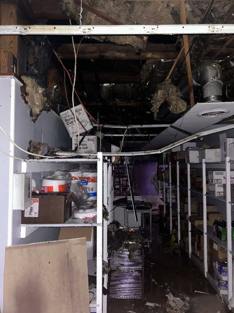 Storage room with shelves, ceiling partially collapsed, insulation and debris scattered, exposed wires and ductwork, damaged boxes and supplies, darkened area indicating possible water or fire damage.