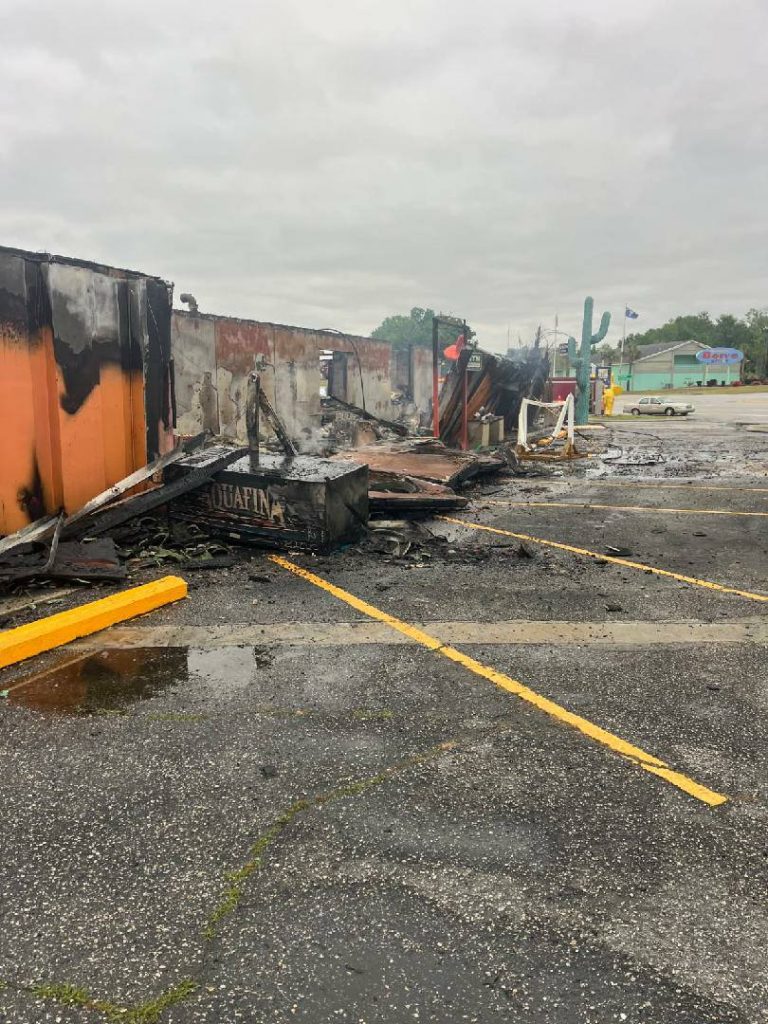 Burnt remains of a building in a parking lot, with charred debris and smoke under a cloudy sky.