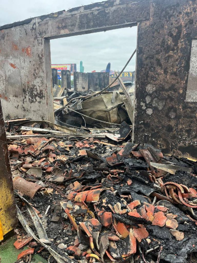 Burnt building with charred walls and debris, including broken tiles and metal, scattered on the ground; visible damage from a recent fire.